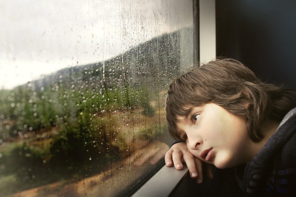 Imagem de um menino triste e debruçado no para peito de uma janela de vidro. A janela está fechada e nela constam pingos de chuva.
