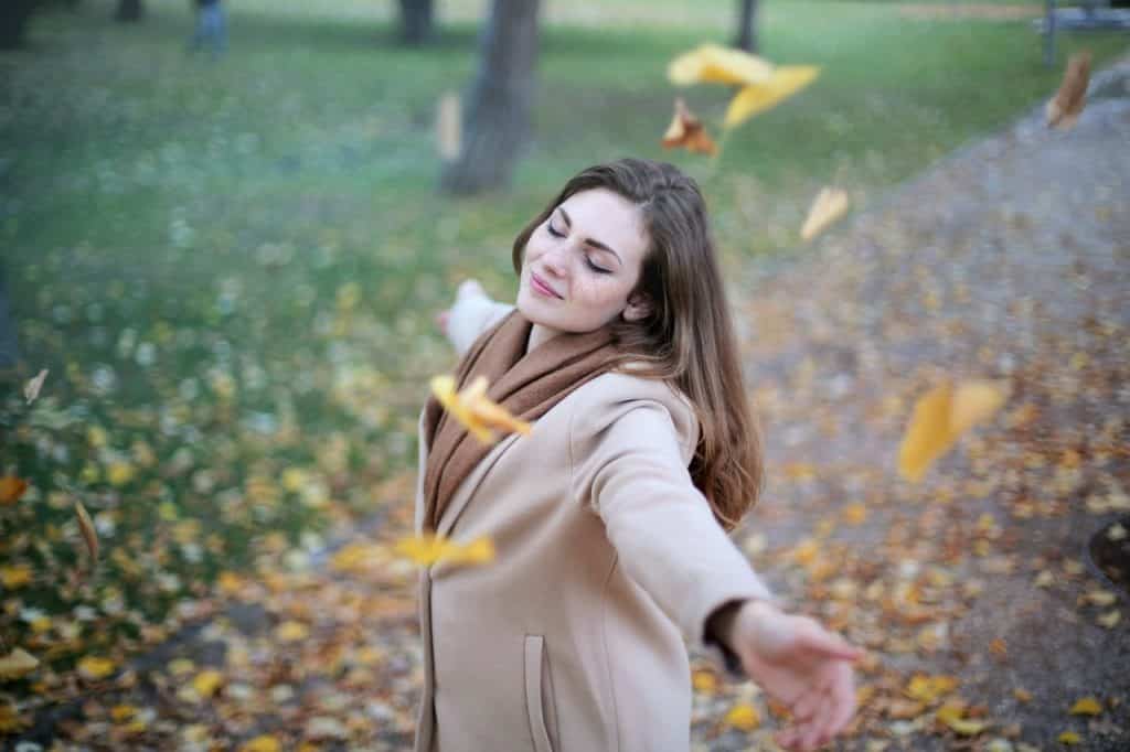 Mulher branca com roupas de frio e braços abertos num parque.