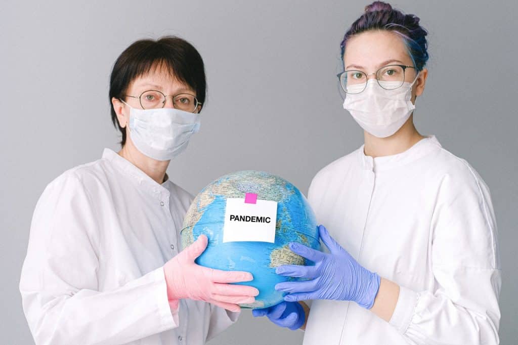 Duas mulheres de máscara e roupa branca segurando o globo com uma plaquinha de "pandemia"