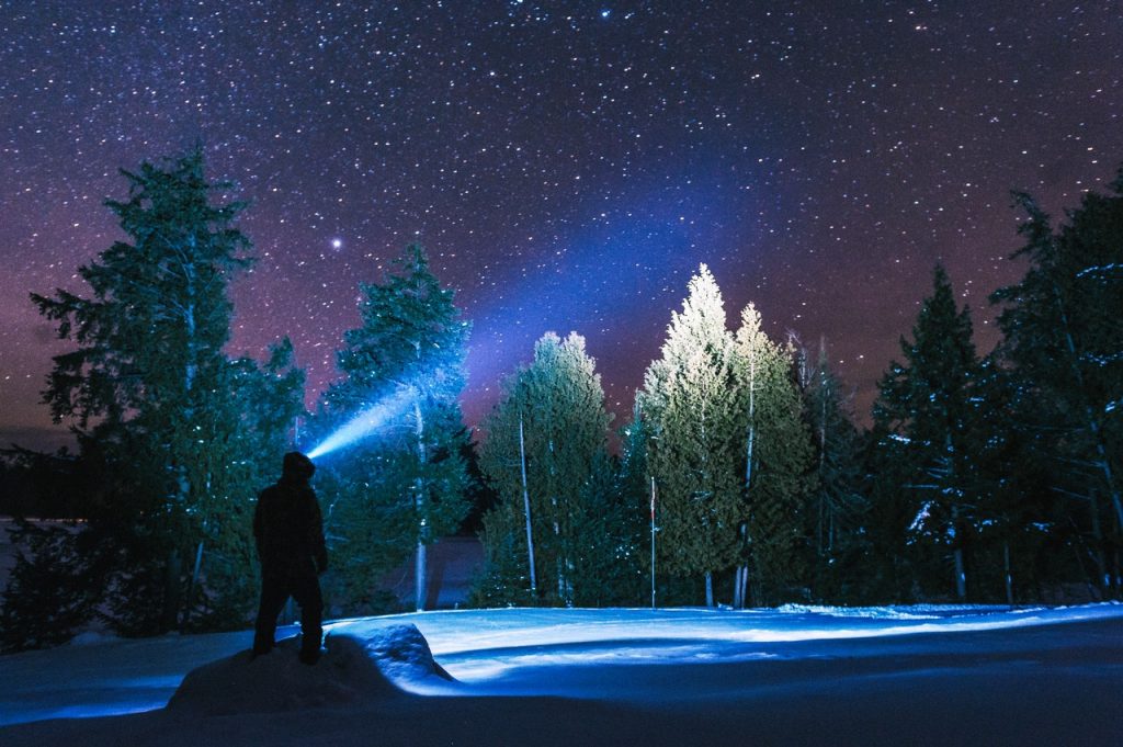 Silhueta de um homem segurando uma lanterna apontada para o alto. Está uma noite estrelada em montanhas cheias de neve.