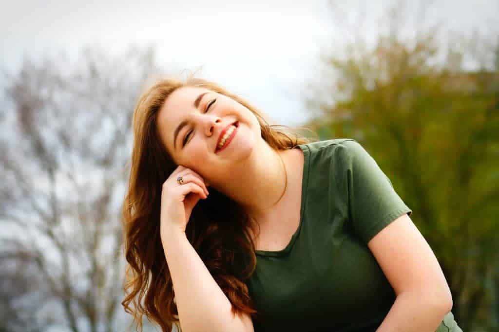 Mulher com blusa verde sentada no chão com os olhos fechados e uma das mãos apoiada na cabeça enquanto sorri. 