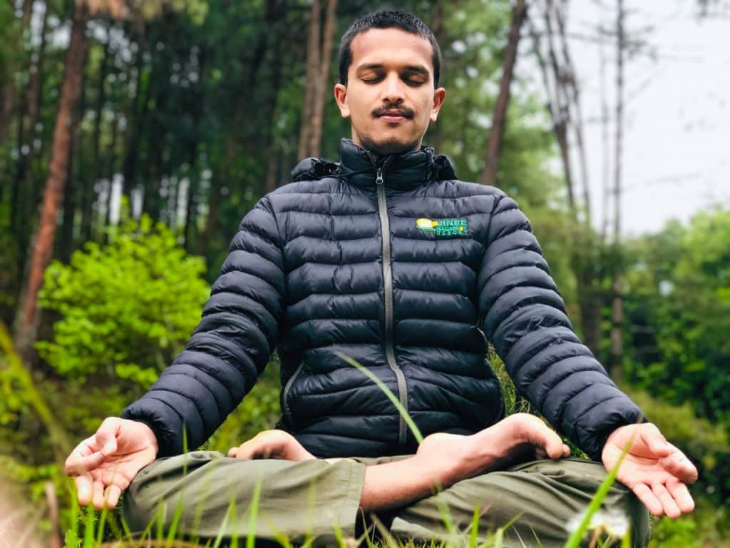Homem de jaqueta azul e calças-verde meditando no meio da natureza.