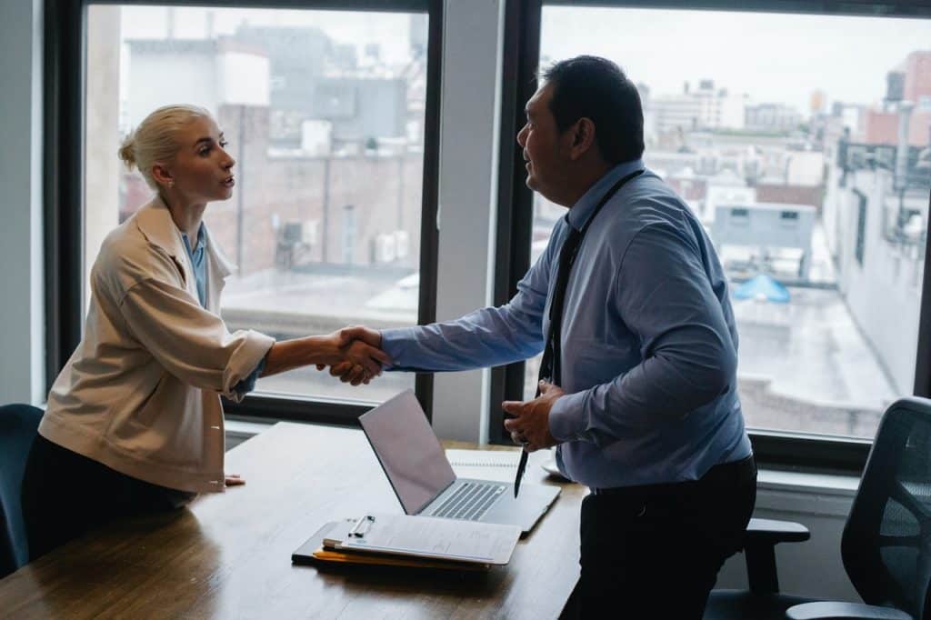 Homem e mulher de roupas sociais apertando as mãos dentro de um escritório.