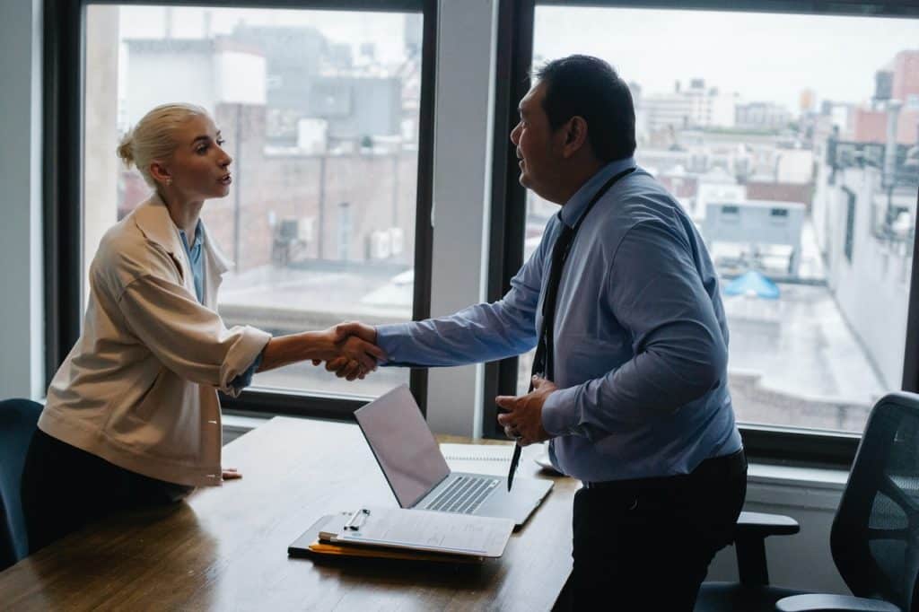 Homem e mulher de roupas sociais apertando as mãos dentro de um escritório. 