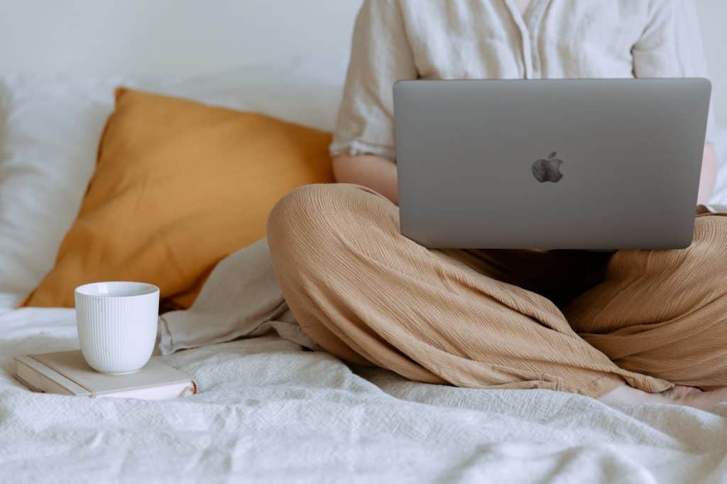 Recorte de uma mulher sentada na cama com o notebook no colo e com uma xícara de café ao lado.