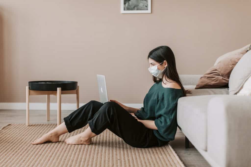 Menina de máscara descartável sentada no chão da sala mexendo no notebook. 