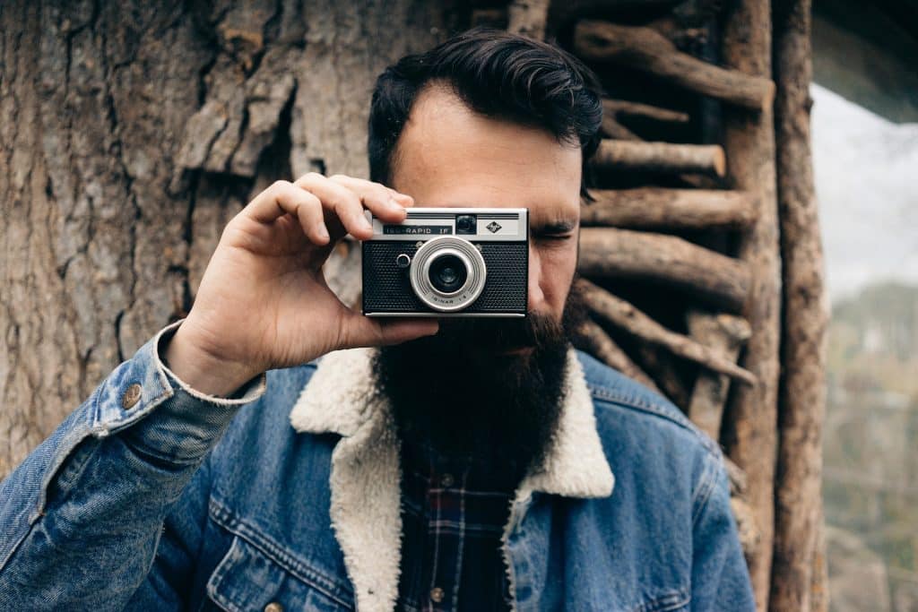 Imagem de um jovem de cabelos escuros, barba e bigode. Ele está encostado em uma árvore, usa uma jaqueta jeans com pele de carneiro e segura em uma das suas mãos uma máquina fotográfica.