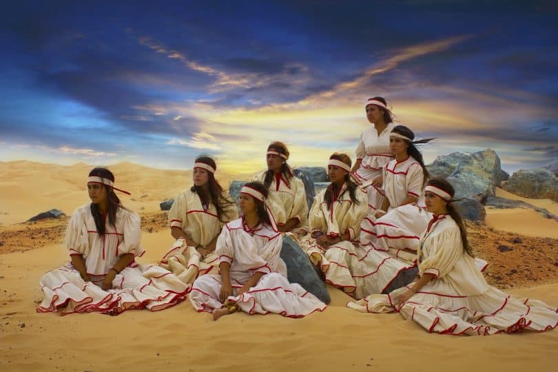 Imagem de oito mulheres indígenas em um terreiro dentro de sua tribo. Elas usam cabelos longos e soltos e uma faixa decorativa na cabeça. Também usam vestidos longos, na cor bege com detalhes na cor vermelha. Todos os vestidos são iguais.