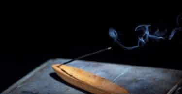 Imagem de fundo preto e em destaque uma mesa de madeira e sobre ela um incenso de citronela sendo queimando. Ele está disposto em um incensário de madeira na cor bege claro.
