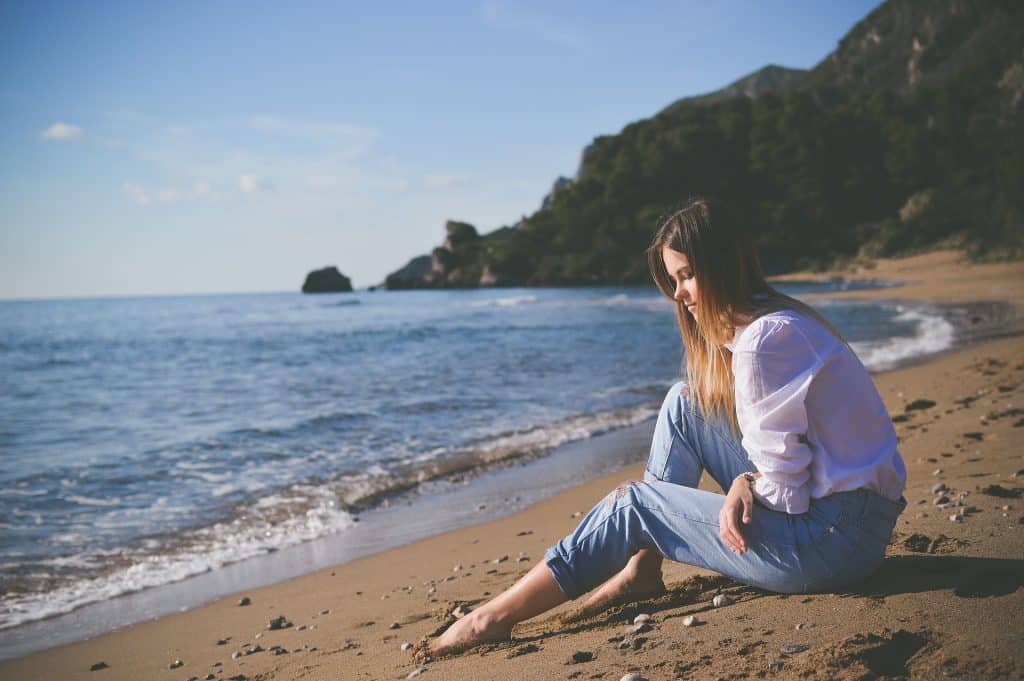 Imagem do mar e em destaque uma mulher de cabelos longos sentada pensativa na areia. Ela está usando calça jeans clara e uma camisa de manga longa na cor branca. Ela está triste e pensativa.
