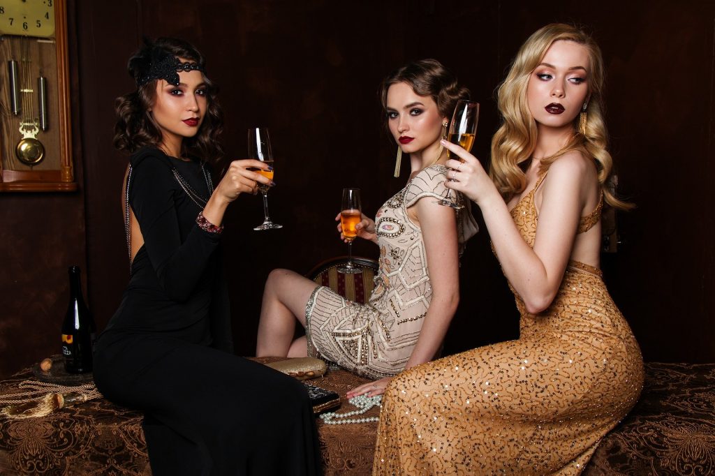 Imagem de três mulheres vestidas para uma festa e cada uma usa um modelo e cor de vestido diferente. As cores são preto, bege e dourado. Todas elas seguram uma taça com bebida e todas estão lindas e bem maquiadas e penteadas.