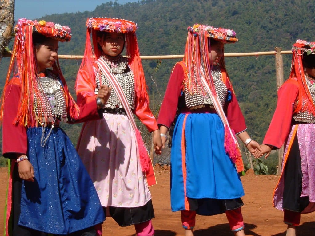 Imagem de quatro mulheres indígenas vestindo roupas coloridas e chapéus ornamentados com fitas e flores. Elas estão de mãos dadas em círculo, prontas para um dança no terreno da tribo.
