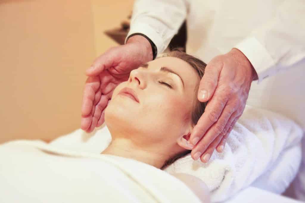 Imagem de uma mulher deitada em uma maca revistada com uma toalha branca. Ela está recebendo de um homem o reiki. Ele está com as mãos levemente impostas sobre os ouvidos da paciente.