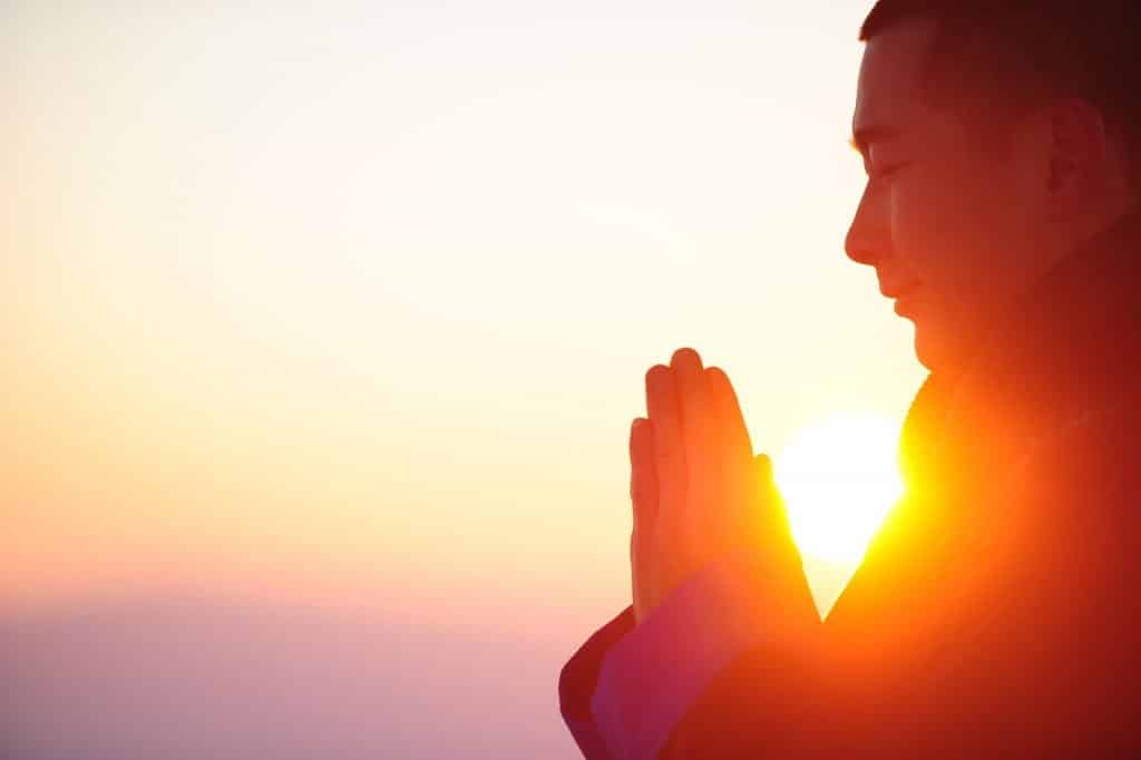 Imagem com o fundo do por do sol e em destaque um homem de perfil com as mãos unidas fazendo meditação.