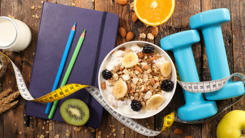 Foto representativa de uma dieta com banana e aveia em um pote ao lado de pesos, fita métrica e lápis