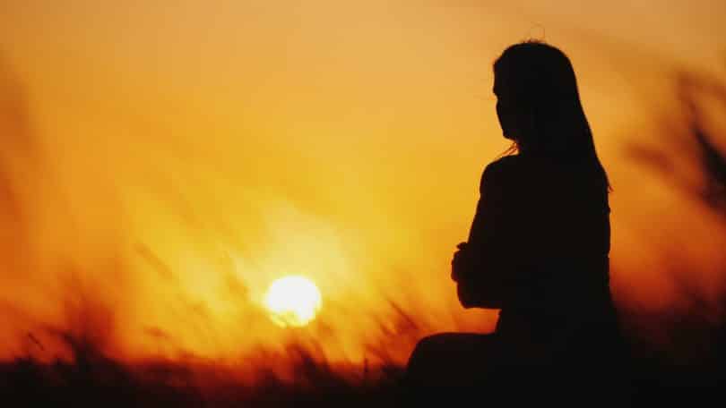 Silhueta de mulher sentada em grama ao pôr do sol.