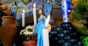 Imagem de um lindo altar com várias estátuas pequenas de Iemanjá, algumas velas brancas acesas e um vaso de flor branca.