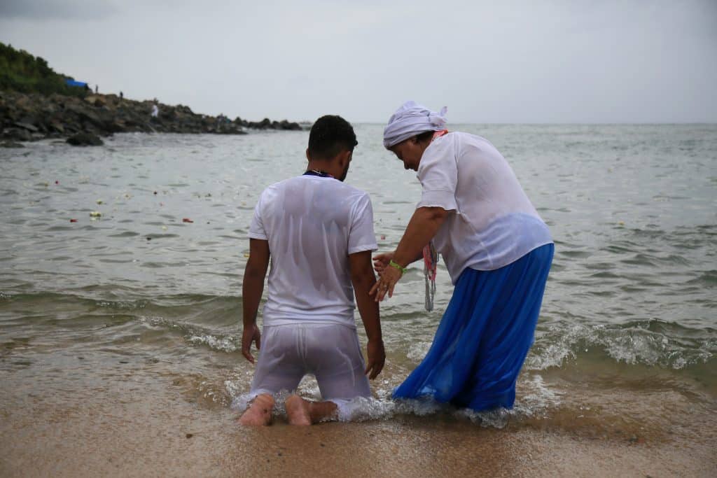 Imagem de um filho de santo, ajoelhando de frente para o mar, se banhando com as águas e se purificando com o banho de Iemanjá. Ao lado dele, sua mãe de santo. Ela usa saia azul longa, camisa branca e um turbante na cor branca na cabeça.
