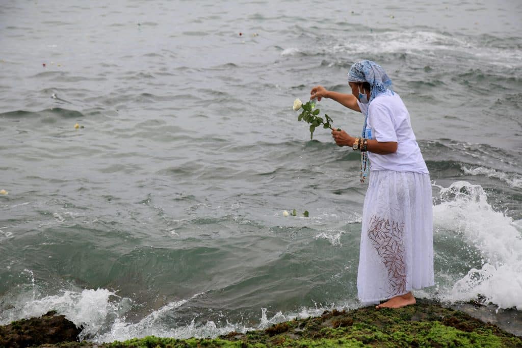 Imagem do mar e de uma mãe de santo jogando pétalas de rosas-brancas na água preparando o banho de Iemanjá.