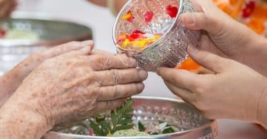 Imagem de um benzimento nas mãos de uma senhora. A benzedeira está lavando as mãos dela com água aromatizada que está dentro de um pote prateado.