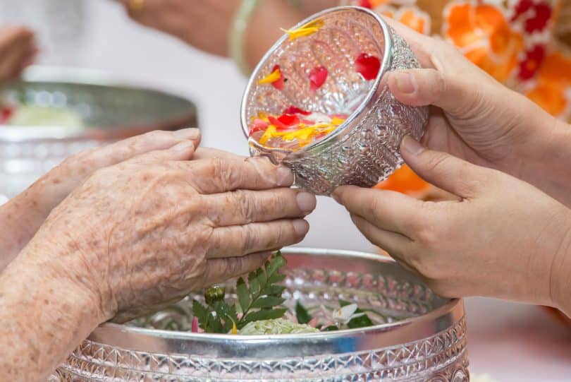 Imagem de um benzimento nas mãos de uma senhora. A benzedeira está lavando as mãos dela com água aromatizada que está dentro de um pote prateado.