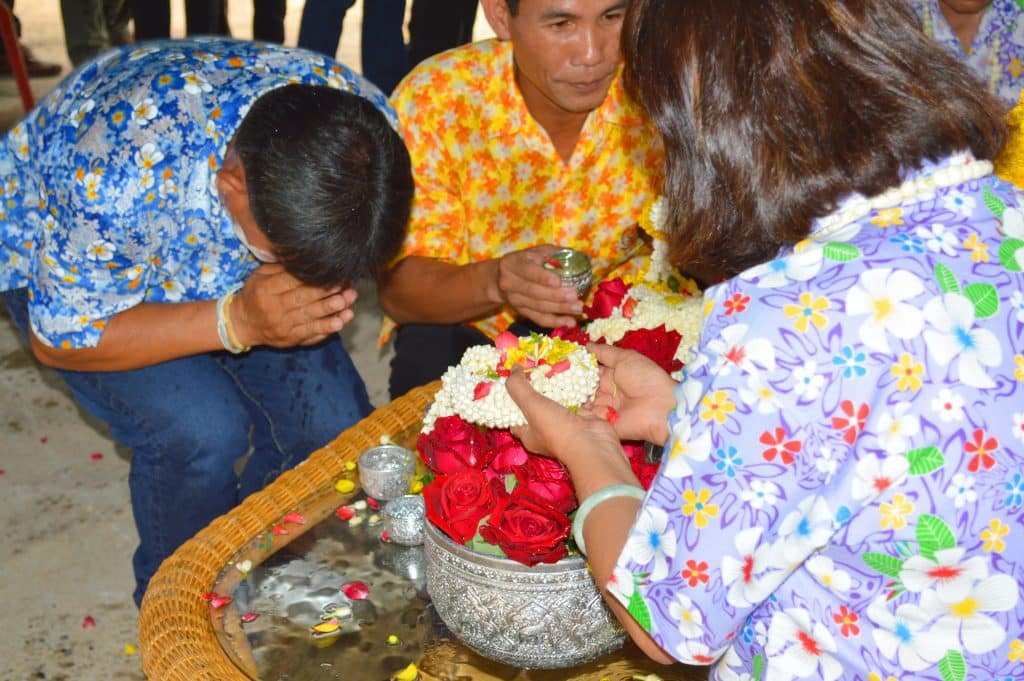 Imagem de dois homens sendo benzidos por uma senhora em um ritual onde ela coloca várias flores e água.
