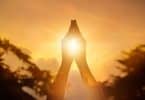 Recorte de uma mão orando com o pôr do sol ao fundo.