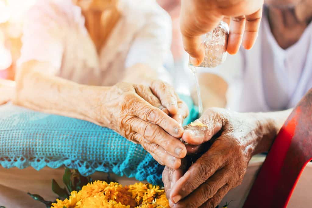 Imagem das mãos de um casal de idosos recebendo o benzimento com água. As mãos dela está disposta sobre uma almofada de crochê azul.
