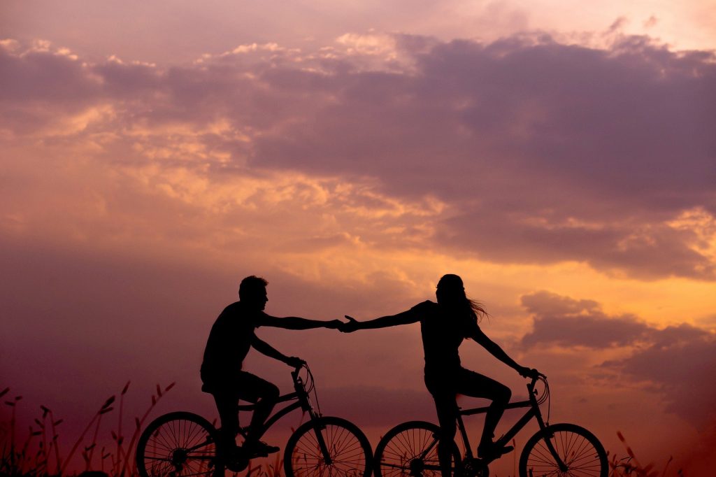 Imagem de um lindo pôr do sol e em destaque um casal sobre suas bicicletas um dando a mão para o outro.
