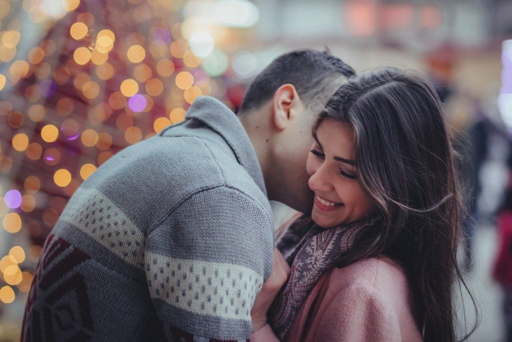 Imagem de um jovem casal e ao fundo algumas luzes amarelas. Ele abraça a mulher que sorri e está muito feliz. Ambos usam blusas de lã, pois está frio.
