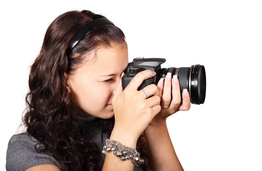 Imagem de uma linda mulher de cabelos longos e cacheados segurando uma câmera fotográfica digital em suas mãos e pronta para fotografar.
