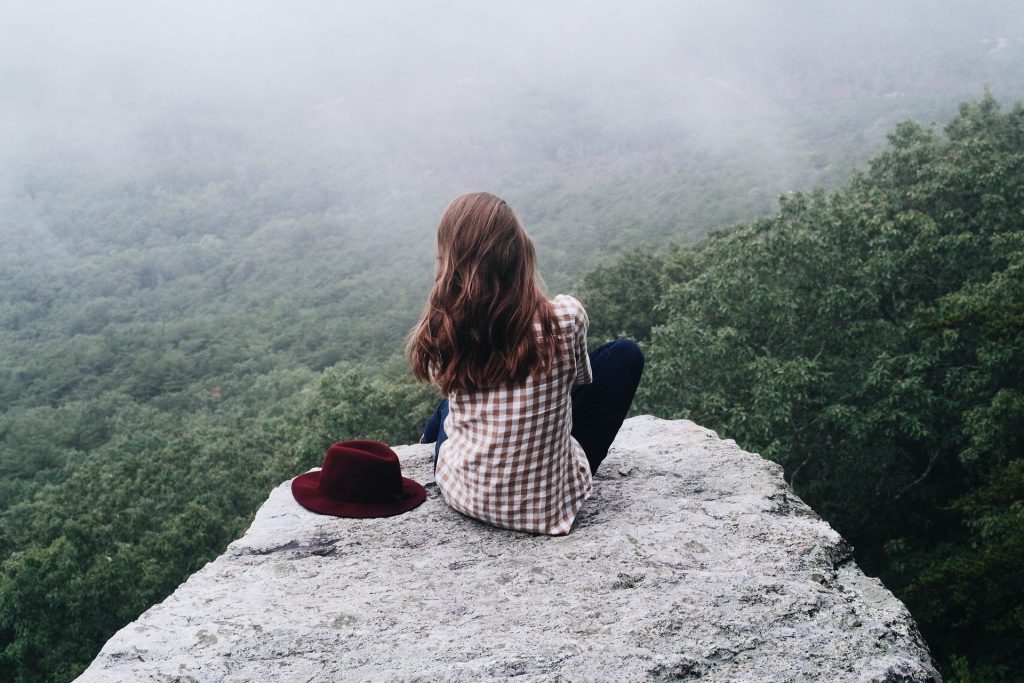 Imagem de uma jovem sentada em uma pedra. Ela usa camisa xadrez nas cores vermelho e branco, calças jeans escura e ao lado dela um chapéu na cor vermelha. Ao fundo uma grande mata encoberta com muitas árvores.