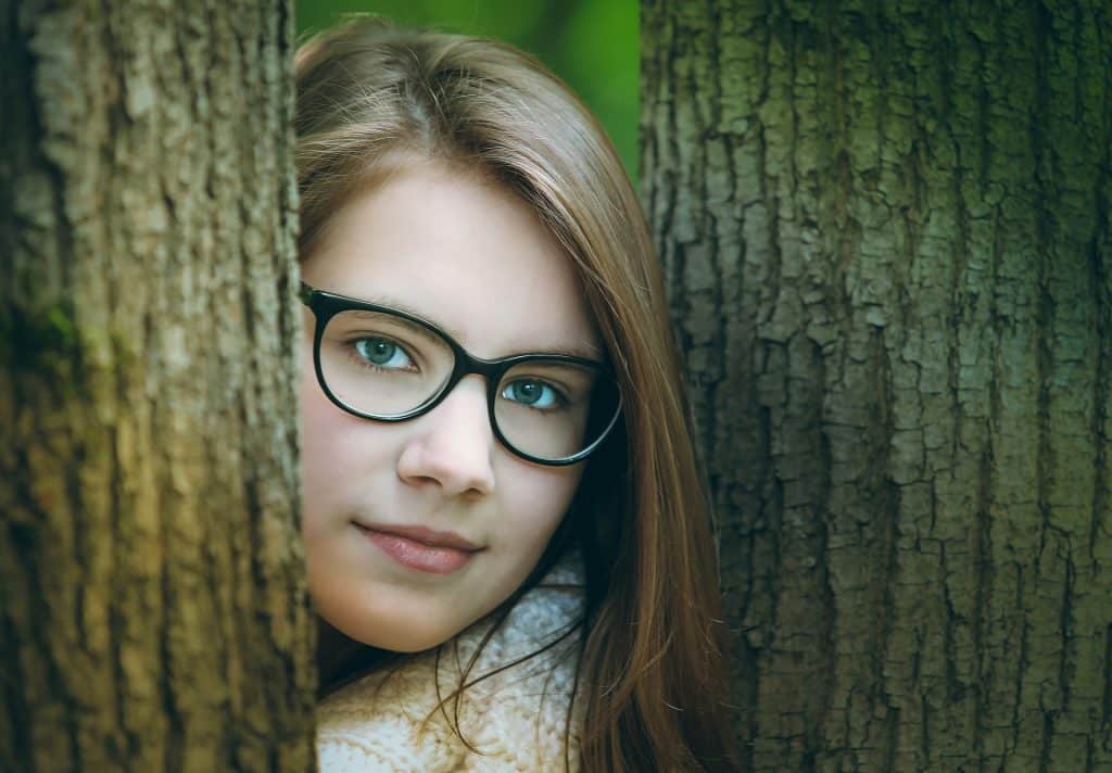 Imagem do rosto de uma jovem entre duas árvores. Ela usa o cabelo solto e um óculos de grau.