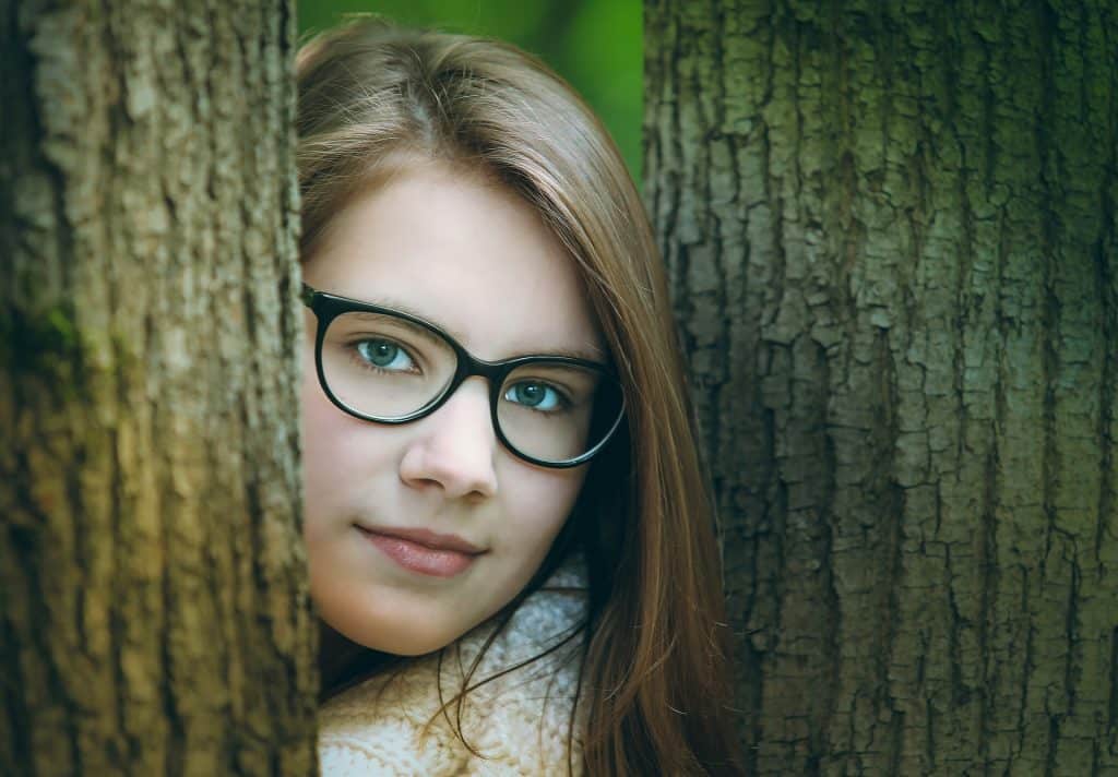 Imagem do rosto de uma jovem entre duas árvores. Ela usa o cabelo solto e um óculos de grau.
