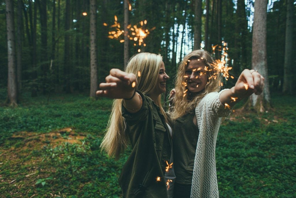 Imagem de duas amigas em uma floresta. Elas estão sorrindo e felizes. Ambas são loiras, com cabelos longos e cada uma segura em suas mãos uma vela que solta faíscas. Elas estão com blusas de frio - uma usa jaqueta na cor verde e a outra um cardigan de linha na cor bege.
