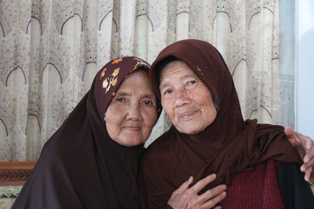 Imagem de duas senhoras sentadas usando lenços sobre suas cabeças. Ambas estão abraças e sendo generosas uma com a outra.