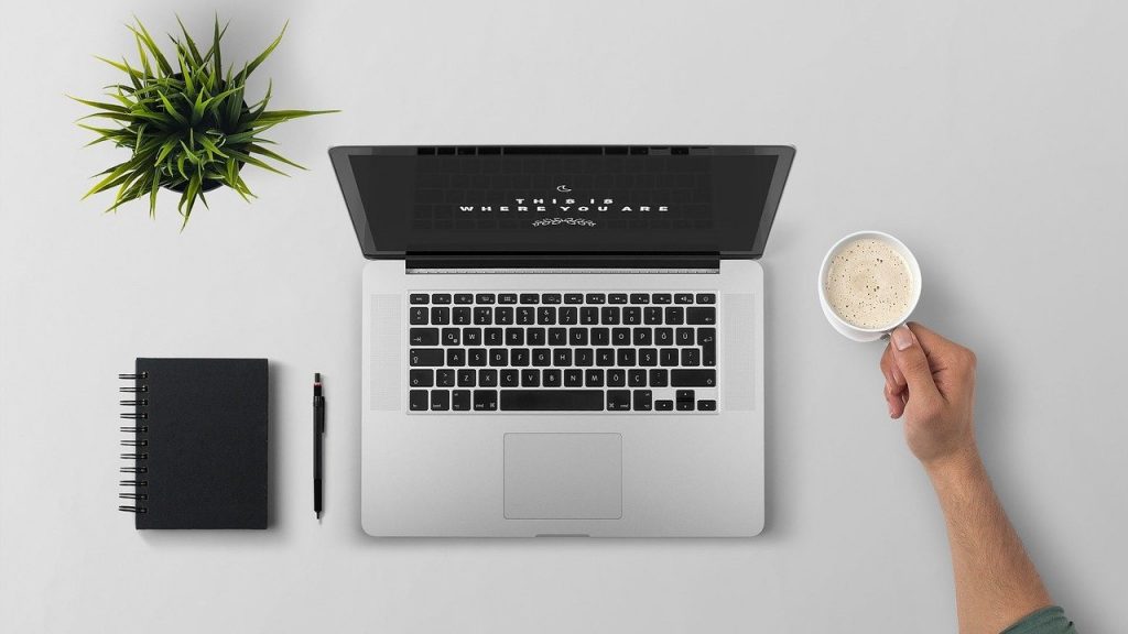 Imagem de uma mesa de trabalho super organizada e limpa. Sobre ela um pequeno vaso de planta, uma agenda e uma caneta na cor preta,  um notebook e a mão de um homem segurando um caneca branca com leite e café.