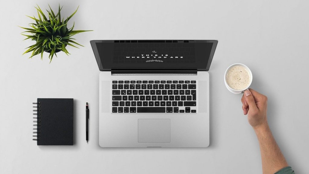 Imagem de uma mesa de trabalho super organizada e limpa. Sobre ela um pequeno vaso de planta, uma agenda e uma caneta na cor preta,  um notebook e a mão de um homem segurando um caneca branca com leite e café.
