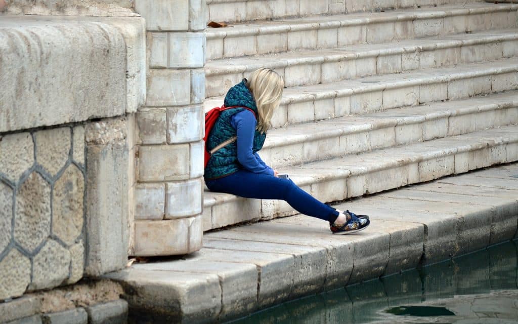 Imagem de uma jovem gatora de cabelos loiros, toda agasalhada e com uma mochila vermelha nas costas. Ela está sentada em uma grande escadaria. Está cabisbaixa e se sentindo culpada por ter feito algo.