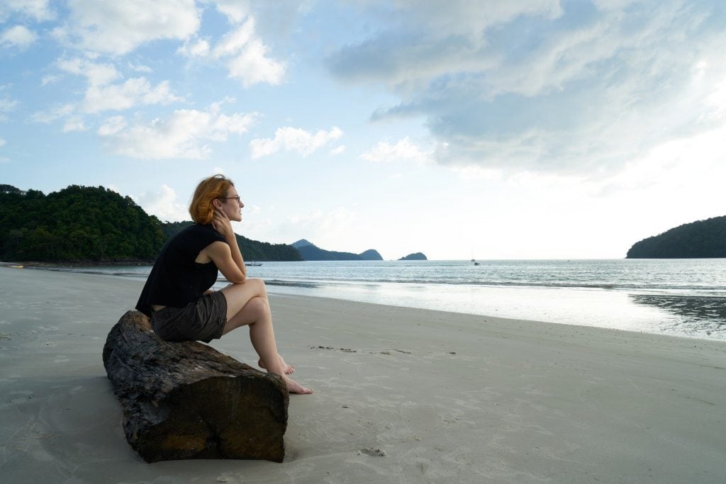 Imagem de uma mulher solitária sentada em um tronco disposto na areia de uma praia deserta. Ela usa uma bermuda e uma camiseta azul e está olhando para o mar.