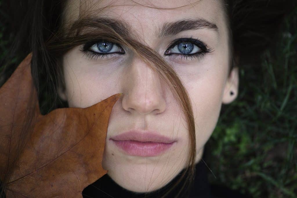 Imagem do rosto de uma modelo de pele branca, olhos azuis e lábios cor de rosa. Ela está deitada sobre um gramado e sobre o rosto dela uma folha de outono na cor marrom.
