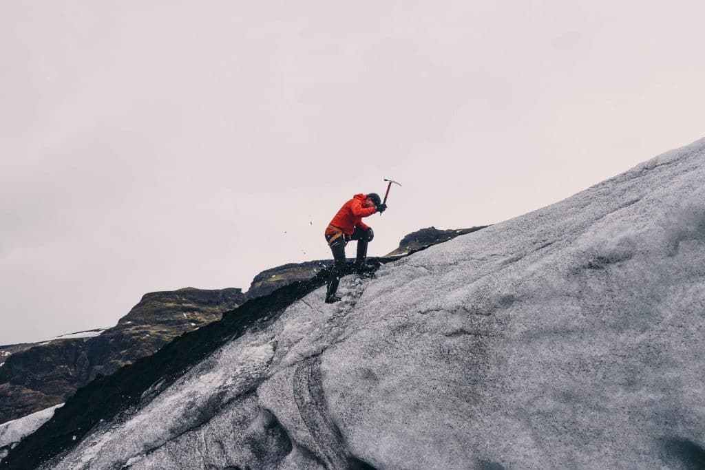 Imagem de um homem vestido com roupa apropriada para o inverno rigoroso. Ele está escalando uma montanha coberta com neve. Em uma das mãos ele segura uma picareta.