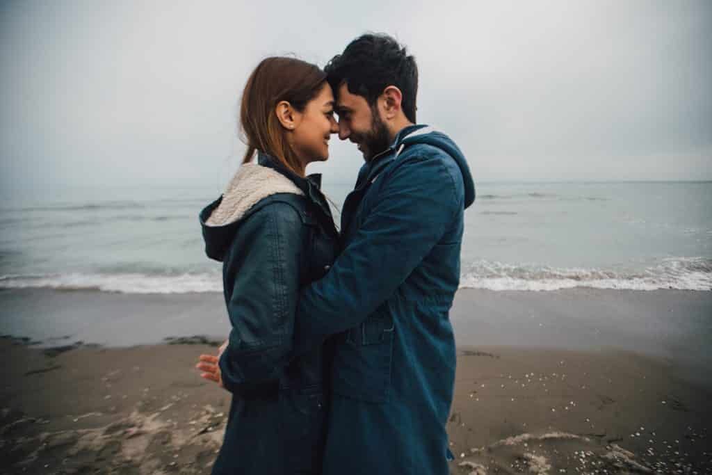Imagem de fundo do mar no inverno e em destaque um jovem casal se abraçando na frente do mar. Ambos usam uma jaqueta de frio com touca.