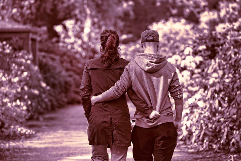 Imagem preta e branco de um casal de costas caminhando abraçados por uma trilha arborizada. Ambos usam roupas de inverno.
