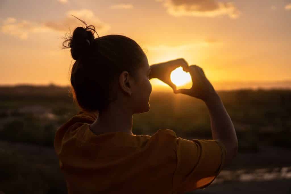 Mulher faz formato de coração com as mãos. Ao fundo, há o pôr do sol. 