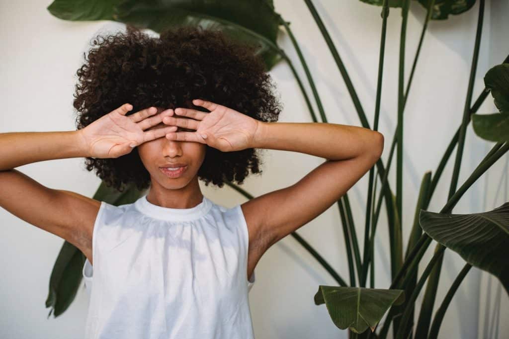 Mulher com as mãos sobre os olhos. Ao fundo, há algumas plantas.