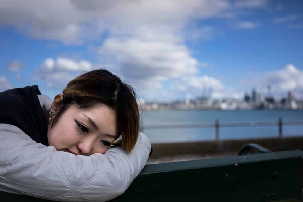 Imagem de fundo de um lindo lago e em destaque uma mulher japonesa sentada em um banco de madeira. Ela usa uma jaqueta de frio na cor cinza com capuz preto. Ela está com a cabeça debruçada no braço e meio se sentindo culpada por ter feito algo.
