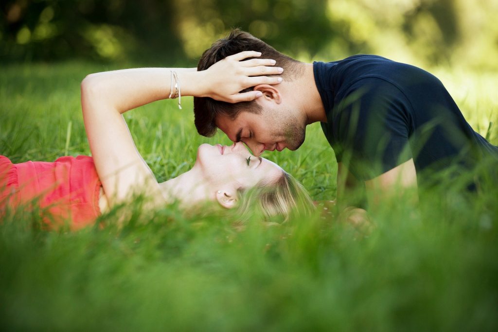 Imagem de um casal se acariciando. Eles estão em um gramado. Ela está deitada e ele abaixado com o rosto sobre o rosto dela.