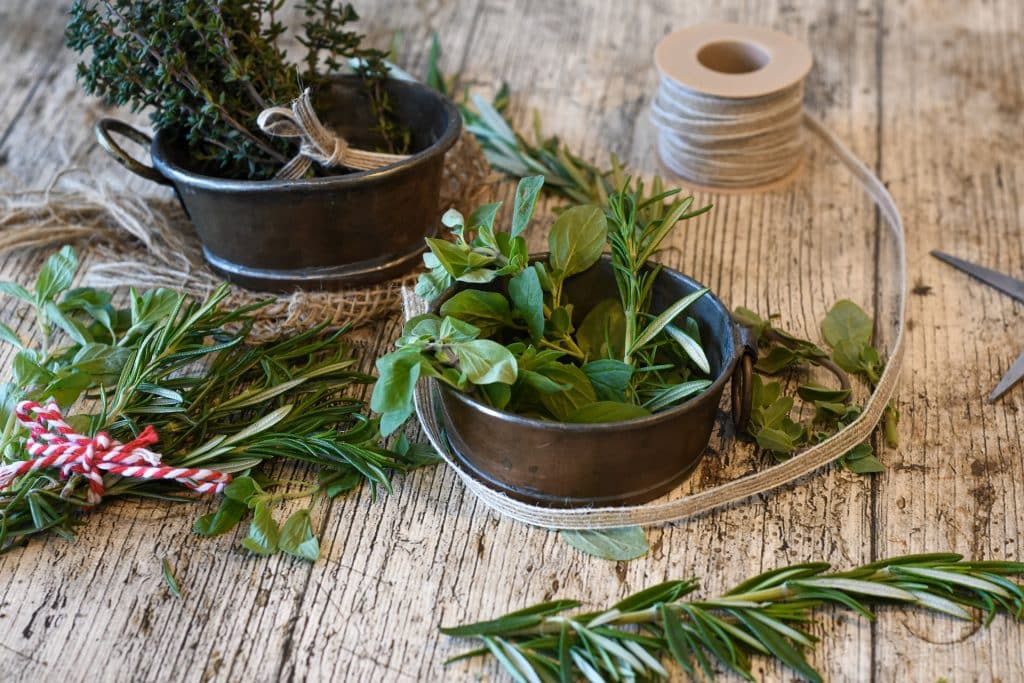 Imagem de vários tipos de ervas dispostos em potes de alumínio e também sobre uma mesa de madeira de fundo branco. Eles estão sendo separados para o preparo de um banho aromático.
