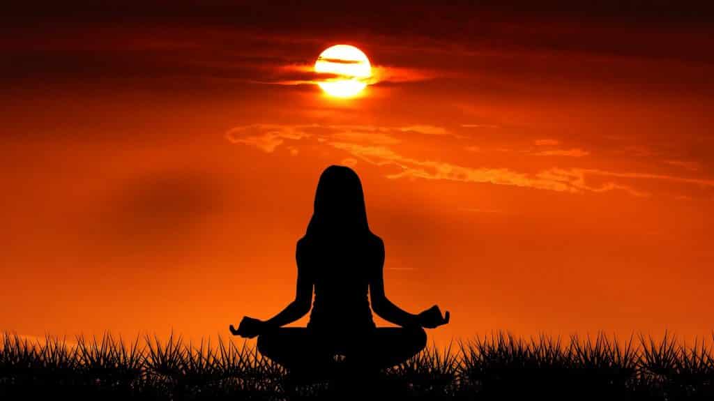 Imagem de fundo avermelhada de um lindo nascer do sol. Em Destaque a silhueta de uma moça sentada de frente para ele, praticando meditação.